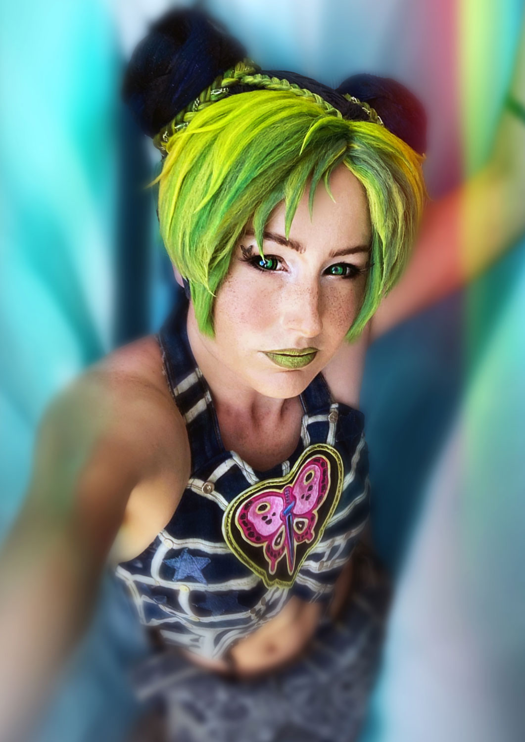 Die Cosplayerin Nomes verkörpert Jolyne mit einer auffälligen Frisur: Der Pony ist in einem leuchtenden Grün gehalten, während die Buns schwarz sind. Ihre grünen Augen strahlen und der grüne Lippenstift ergänzt ihr Gesamtbild. Der Hintergrund ist bunt und leicht verschwommen, was die lebendige Ausstrahlung des Charakters unterstreicht.