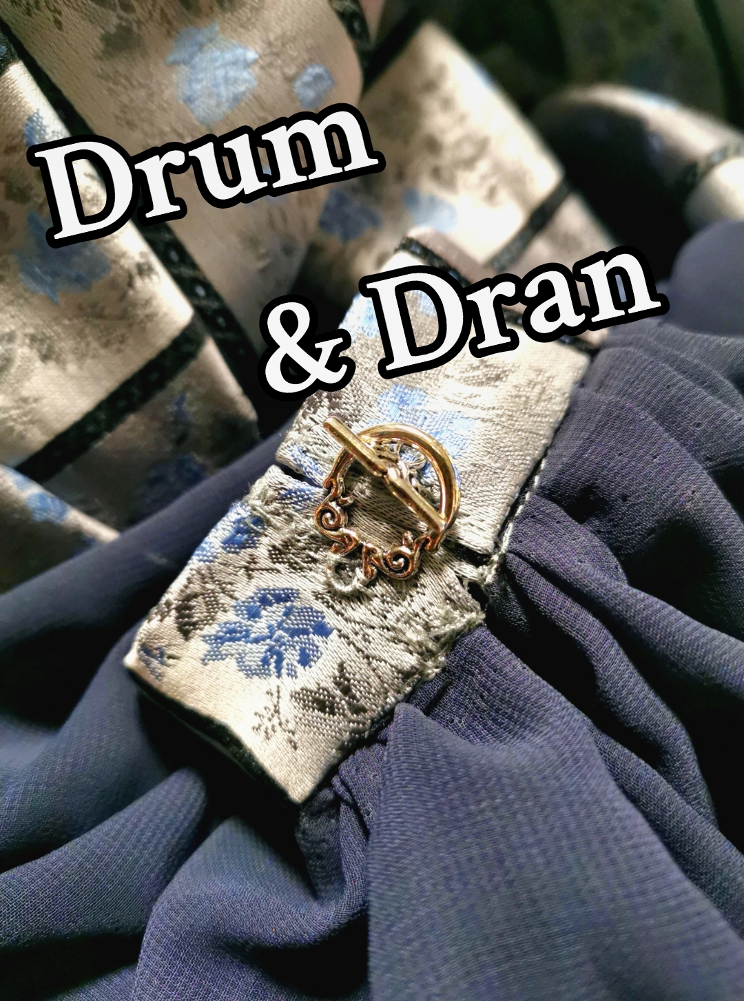 Im Vordergrund steht der Text "Drum und Dran" in weißer Schrift. Im Hintergrund ist ein Ärmel aus blauem Stoff mit einer Silber-Borte und einer Metall-Öse.