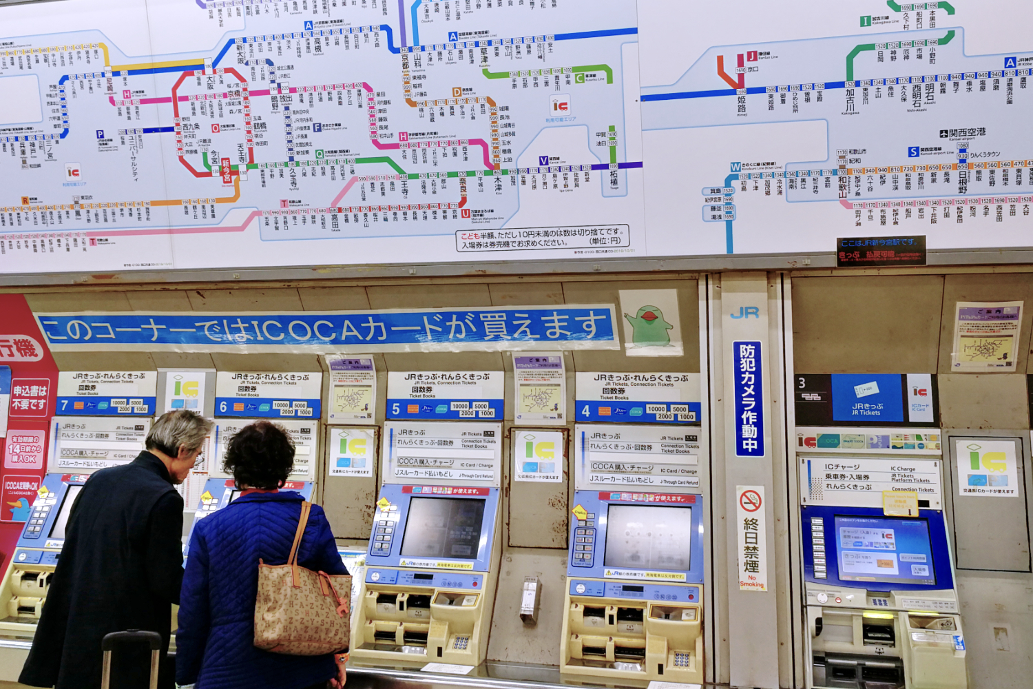 Ein großer Zugfahrplan und ein Ticketautomaten mit zwei Reisenden davor an einem Tokyoter Bahnhof.