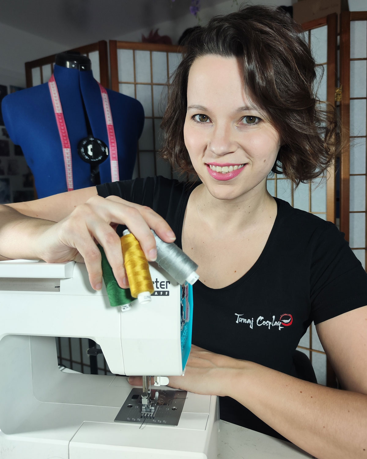 TunajCosplay sitzt vor ihrer Nähmaschine, mit drei Fadenspulen in der Hand. Hinter ihr steht ihre Schneiderpuppe mit einem Maßband um den Hals.
Foto-Credit: TunajCosplay