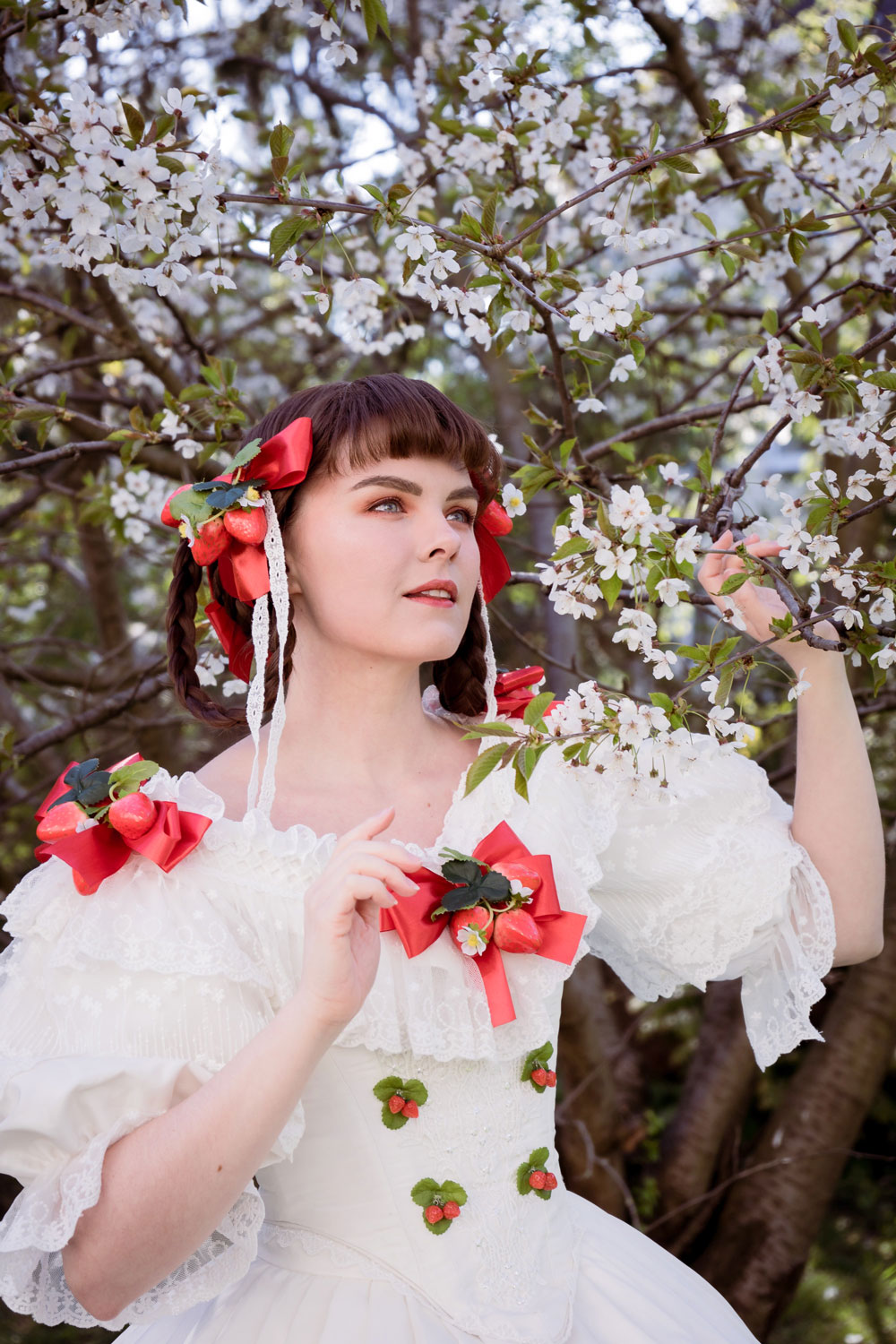Eine Cosplayerin steht zwischen Kirschblütenzweigen und guckt in die Ferne. Sie hat ein weißes spitzenbesetztes Kleid mit schmaler Taille und ausladenden Rock an. Auf dem Kleid befinden sich 3D Erdbeeren.