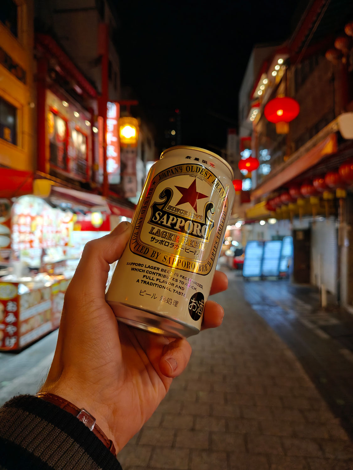 Eine Hand, die ein Sapporo-Bier in den Bildmittelpunkt hält. Im Hintergrund verschwommen eine japanische Einkaufsstraße.