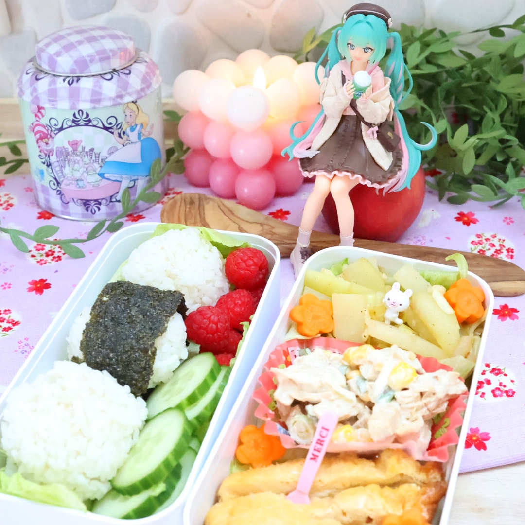 Abgebildet sind zwei Hälften eines Bento, links mit Reisbällchen, Himbeeren und Gurken, rechts eine Mischung aus Frittiertem und Karotten, die in die Form von Blumen geschnitten wurden. Oben links sieht man eine Teedose mit Alice im Wunderland darauf abgebildet, rechts davon, direkt über dem Bento, sitzt eine Hatsune Miku Figur.