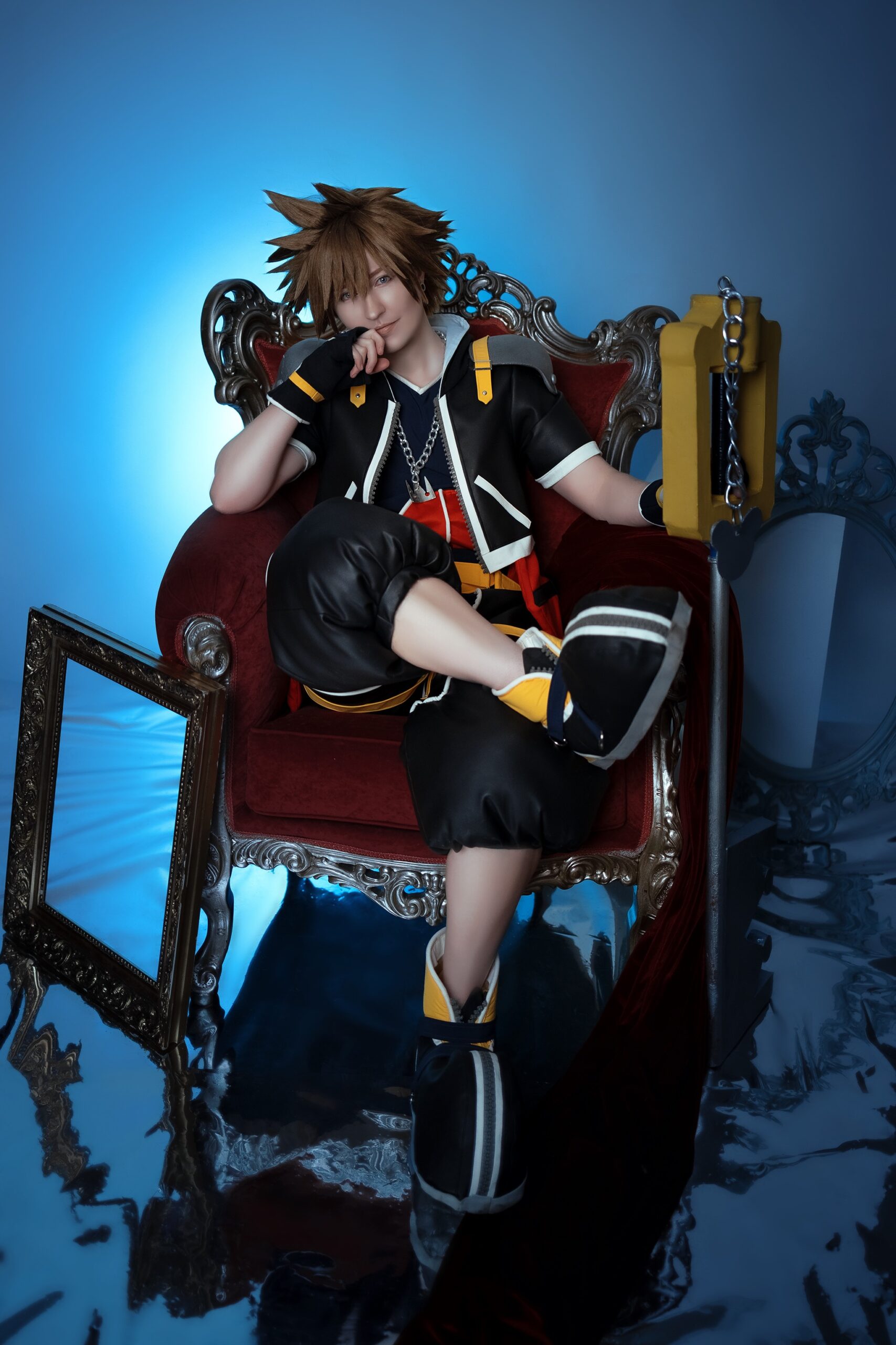 Der Cosplayer Twinfools sitzt in seinem Sora-Kostüm auf einem roten Sessel vor einem blauen Hintergrund, sein Keyblade steht neben ihm.