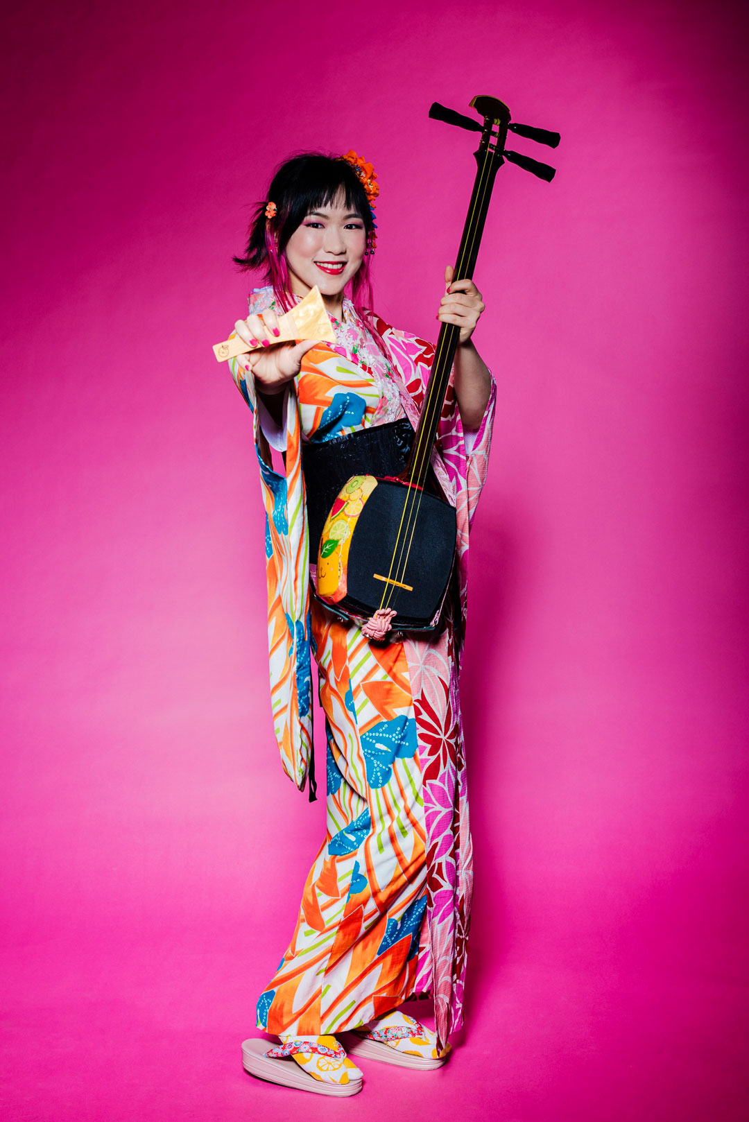 E-Yuzu-HP Die Sängerin Yuzu sitzt vor einem pinken Hintergrund in einem farbgenfrohen Kimono und hält dabei ihr Instrument das Shamisen in der Hand.