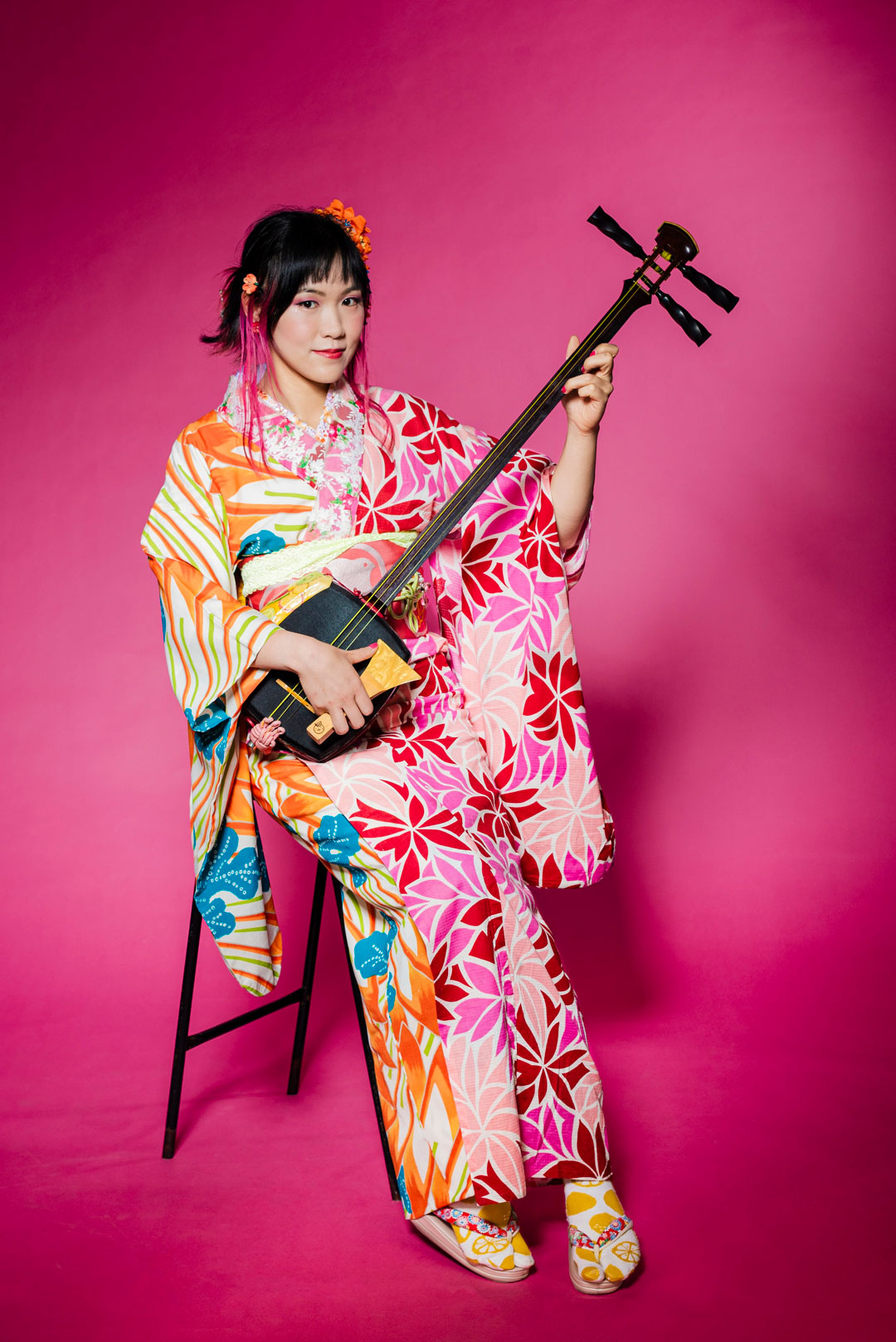 Die Sängerin Yuzu sitzt vor einem pinken Hintergrund in einem farbgenfrohen Kimono und hält dabei ihr Instrument das Shamisen in der Hand.