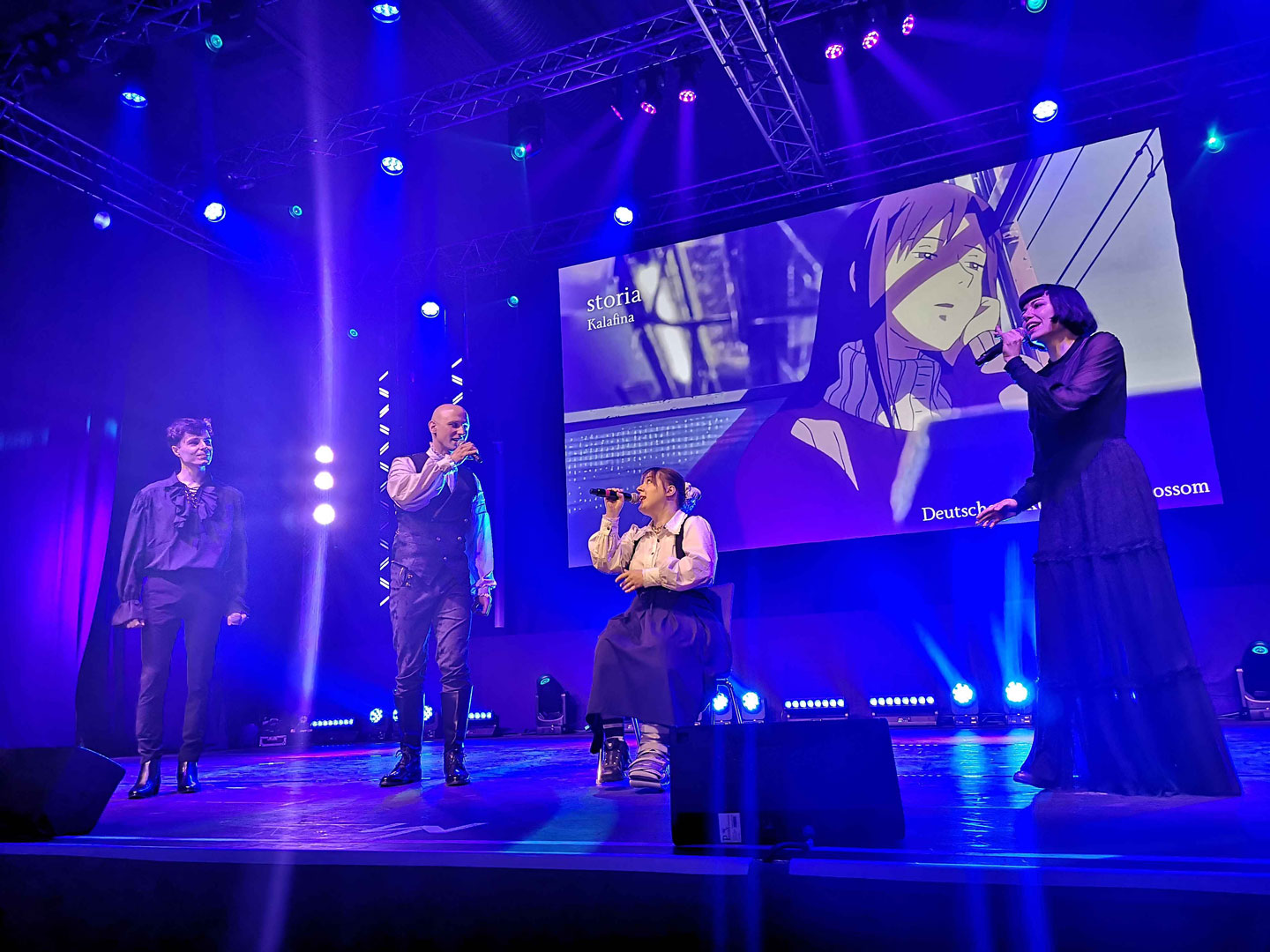 Auf einer in blau und violett ausgeleuchteten Bühne sind die vier Mitglieder von canatamaer bei ihrem Gesangsauftritt zu sehen, das Bild auf dem Backdrop der Bühne zeigt einen Animeausschnitt und den Titel des präsentierten Liedes.