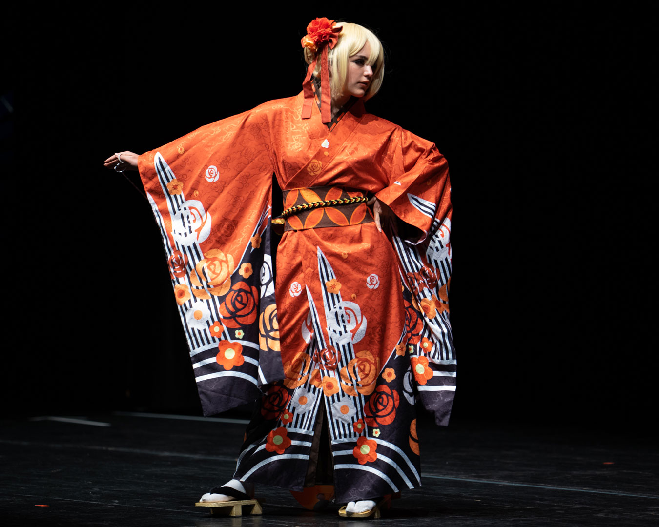 Eine Cosplayerin in starker Pose, welche einen detailreichen japanischen Kimono in orangener Farbe trägt. Auf ihrem Kopf befindet sie eine kurze blonde Perücke und ein farblich zum Kimono abgestimmter Haarschmuck aus Blumen und Bändern. Sie posiert allein auf der Bühne vor schwarzen Hintergrund.