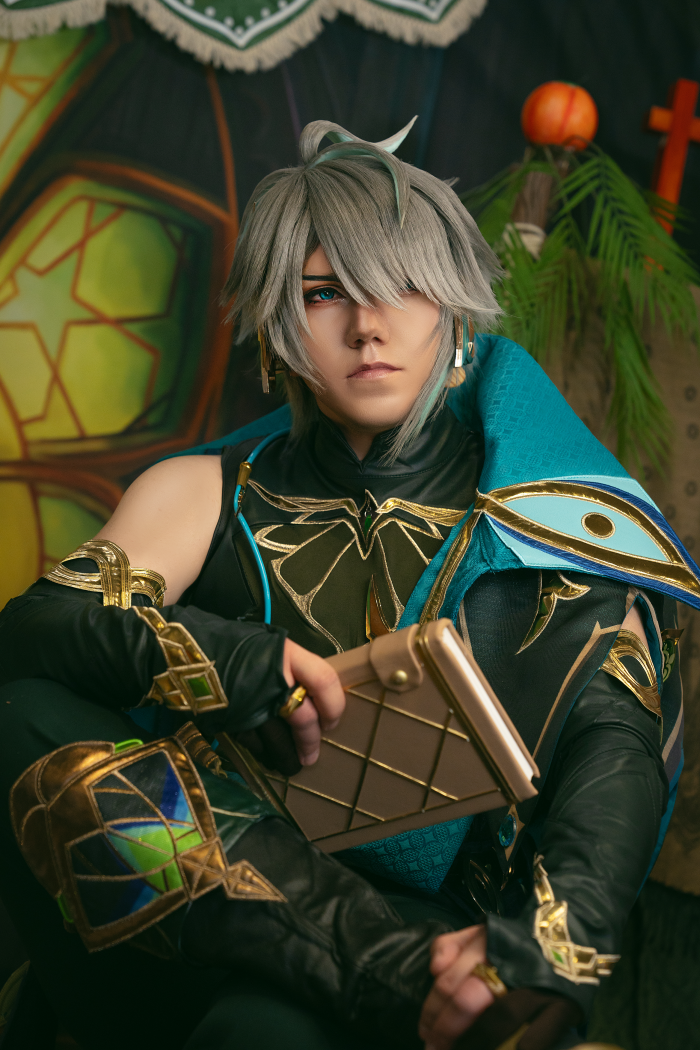 Der Cosplayer Dorian Makbeth sitzt in seinem Alhaitham Kostüm aus Genshin vor einem grünen Hintergrund. Er hält ein Buch in der Hand und trägt eine graue Perücke.