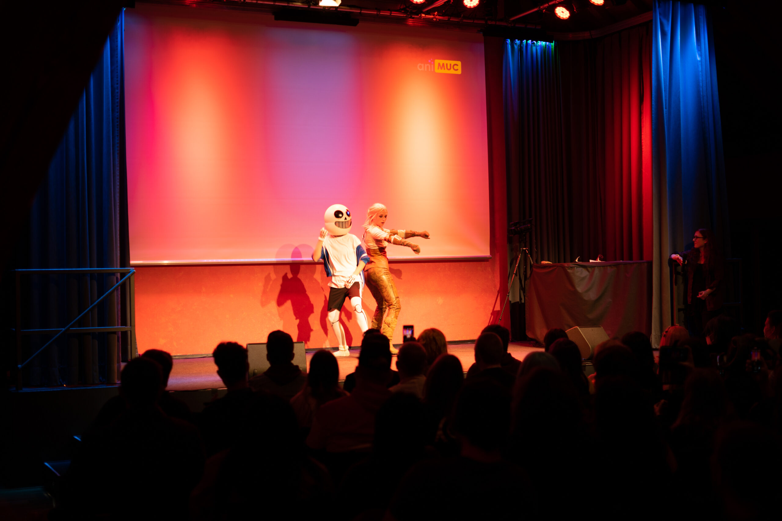 WB-Cosplay Dance-off_ORIGINAL2 Zwei Personen in Kostümen, welche Rücken an Rücken miteinander Tanzen. Die Person auf der rechten Bildseite stellt Sans aus dem Videospiel Undertale dar, die Person auf der linken Bildseite verkörpert Ciri aus der Witcher Videospielreihe.