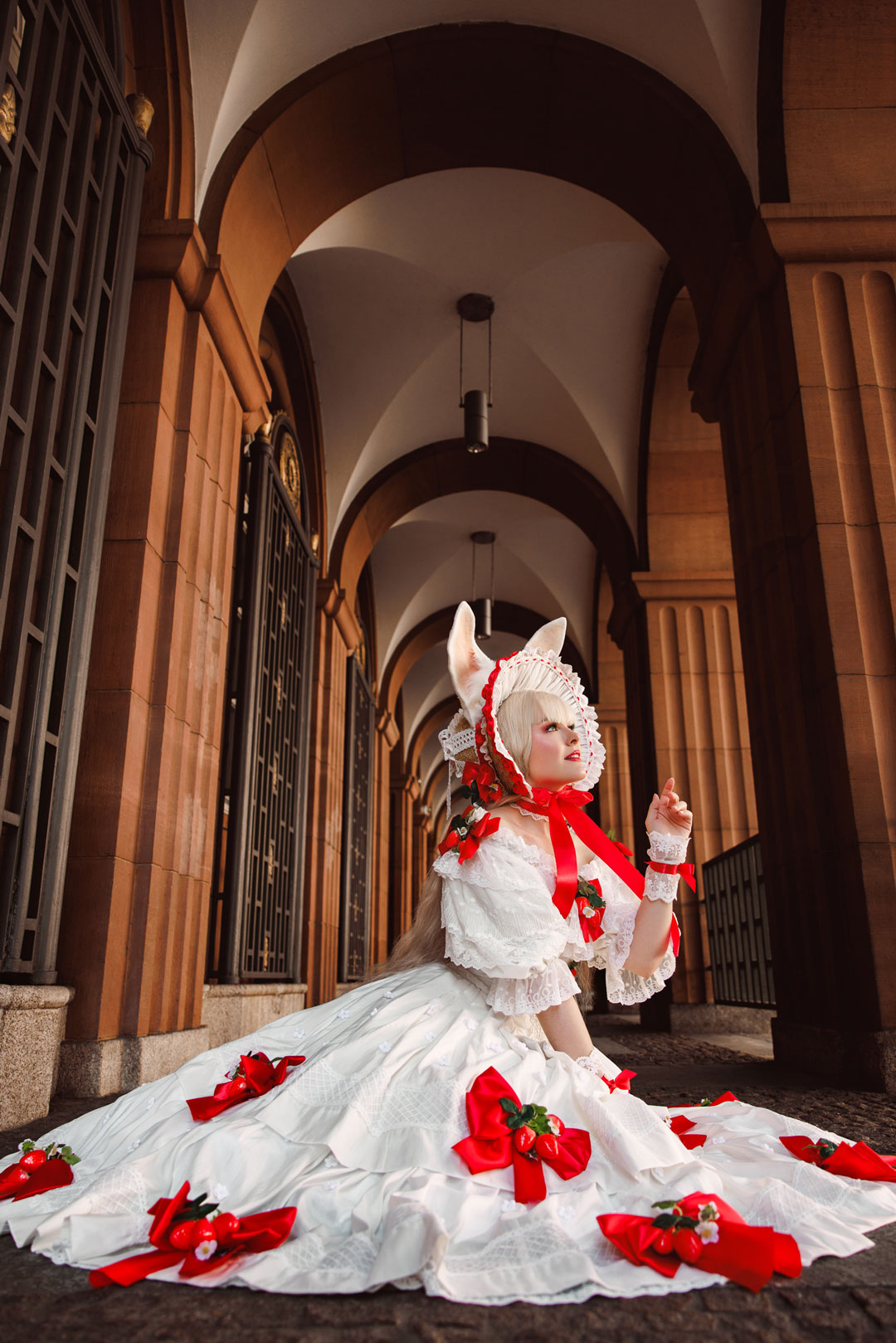 WS-Level-up-your-Cosplay_26WS59-HP Eine Cosplayerin sitzt in einem Gang auf dem Boden. Sie hat eine Hand gehoben und schaut nach oben, als würde sie etwas Schönes sehen. Sie trägt ein weißes reichlich verziertes Kleid und ein Bonnet mit Hasenohren.